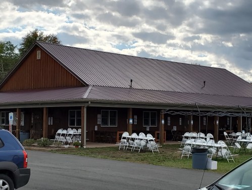 A rustic building with a metal roof, surrounded by chairs set up outside for an event, under a cloudy sky.