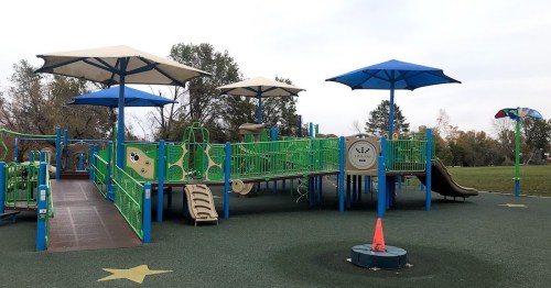 Colorful playground with green structures, slides, and blue umbrellas, set in a grassy park under a cloudy sky.