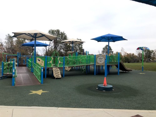 Colorful playground with slides and climbing structures, shaded by blue canopies, set in a grassy park.