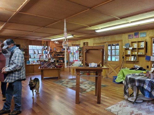 A cozy shop interior with wooden walls, shelves of crafts, a table, and a person with a dog walking by.