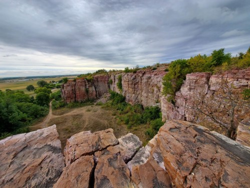 A rocky cliff overlooks a grassy valley under a cloudy sky, with trees scattered around the landscape.