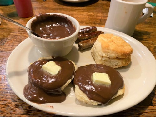 A plate with biscuits topped with chocolate sauce and butter, a bowl of chocolate sauce, sausage links, and a cup.