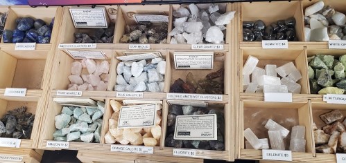 A display of various crystals and gemstones in wooden boxes, labeled with names and prices.