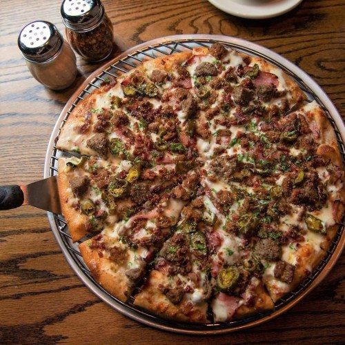 A freshly baked pizza topped with meats, jalapeños, and herbs, served on a wooden table with seasoning shakers nearby.