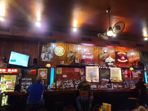 A cozy bar interior with wooden walls, neon signs, and patrons sitting at the counter, surrounded by various liquor bottles.