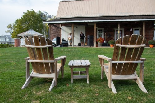 Two wooden Adirondack chairs and a small table face a house with a porch, where people are setting up for an event.