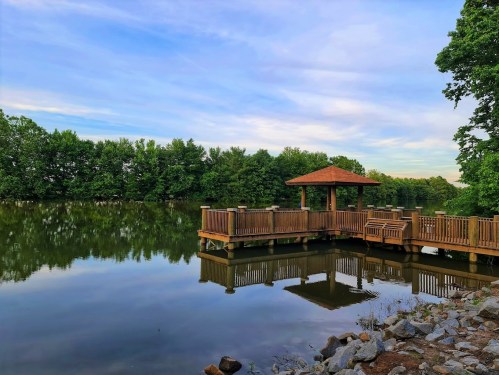 A serene lakeside view featuring a wooden gazebo and reflections on calm water, surrounded by lush greenery.
