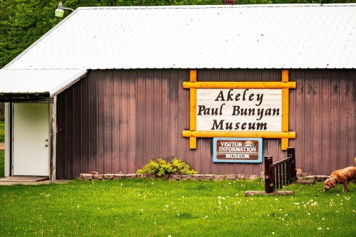 A wooden building with a sign reading "Akeley Paul Bunyan Museum" and a visitor information sign in front.