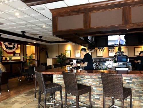 A bar area in a restaurant with a bartender preparing drinks and a TV in the background. Decor includes plants and artwork.