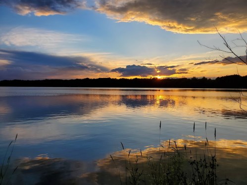 A serene lake at sunset, reflecting vibrant colors in the sky and water, with silhouettes of trees in the background.