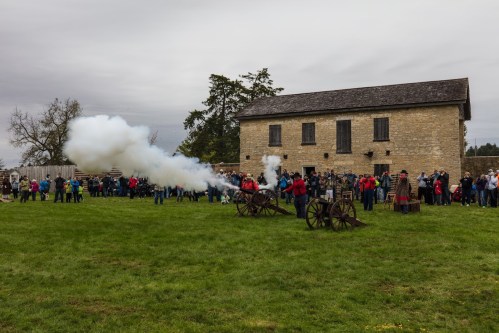 A historic reenactment scene with cannons firing, a crowd watching, and a stone building in the background.