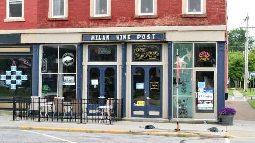 Exterior of a red brick building featuring Milan Wine Post, a yoga studio, and a cafe with outdoor seating.