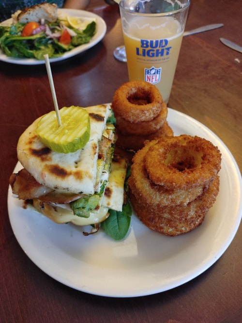 A plate with a stacked sandwich topped with a pickle, alongside onion rings and a glass of beer.