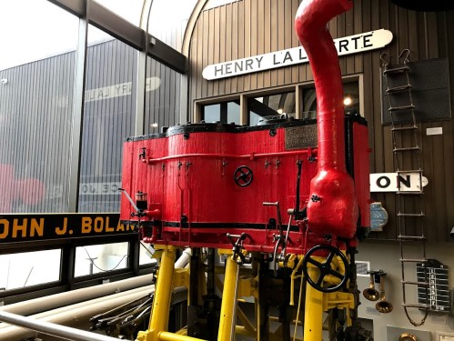 A vintage red steam engine with yellow accents displayed in a museum, featuring various signs in the background.