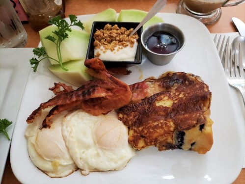 A breakfast plate with two fried eggs, bacon, French toast, yogurt with granola, and melon slices.