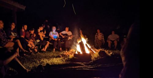 A group of people gathered around a campfire at night, some playing guitar and others enjoying the warmth of the fire.