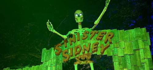 A large skeleton with glowing eyes stands behind a wooden sign that reads "Sinister Sidney," surrounded by green lighting.