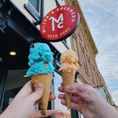 Two hands hold ice cream cones: one with blue ice cream and chocolate chips, the other with orange ice cream, in front of a shop sign.