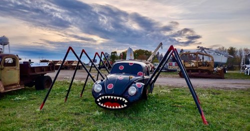 A car transformed into a spider sculpture, featuring a painted face and long legs, set against a cloudy sky.