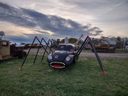 A car transformed into a spider sculpture, with large legs and a painted face, set against a cloudy sky.