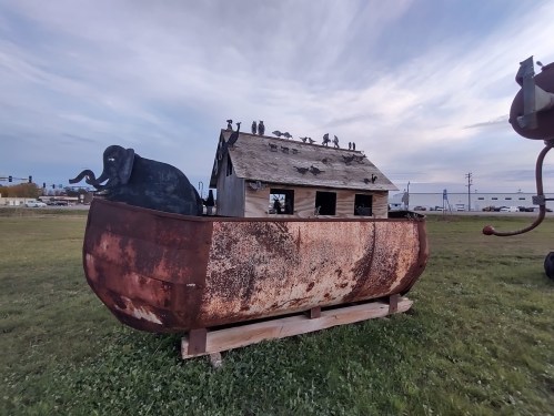 A whimsical sculpture of a house on a boat, featuring an elephant and birds on the roof, set in a grassy area.