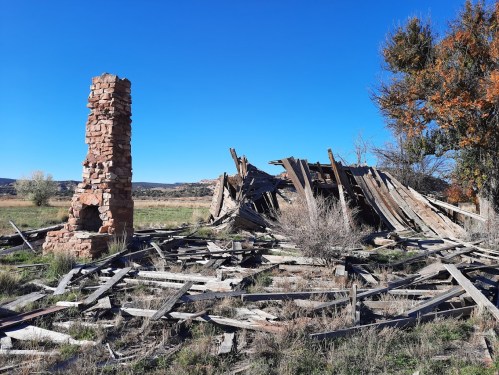 A crumbling stone chimney stands beside a collapsed wooden structure in a grassy field under a clear blue sky.