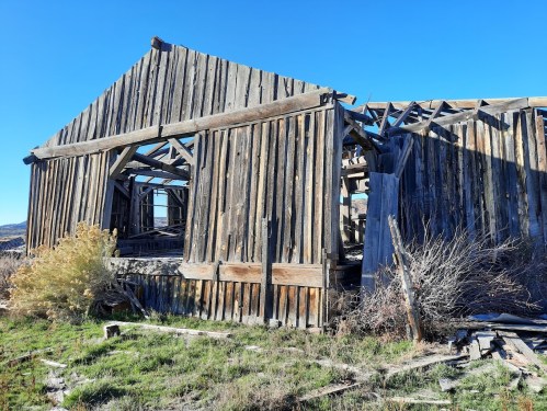 An old, weathered wooden barn with a partially collapsed roof, surrounded by dry grass and shrubs under a clear blue sky.