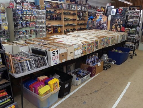A store display featuring comic books, action figures, and collectibles on shelves and tables.