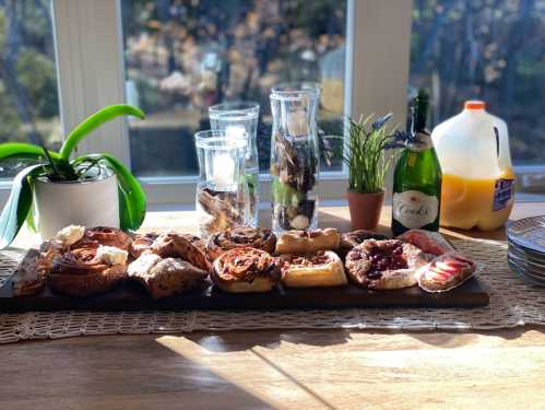 A wooden platter with assorted pastries, drinks, and decorative plants, set by a sunny window.