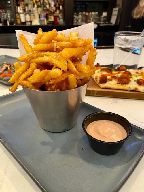 A metal cup filled with golden, crispy fries next to a small container of dipping sauce on a plate.