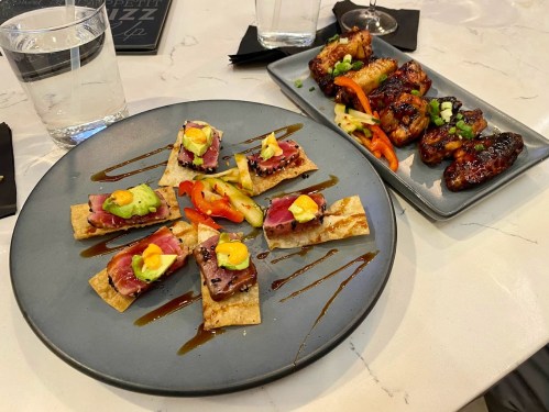 A plate of appetizers featuring small bites with meat, vegetables, and sauces, alongside a dish of glazed chicken wings.