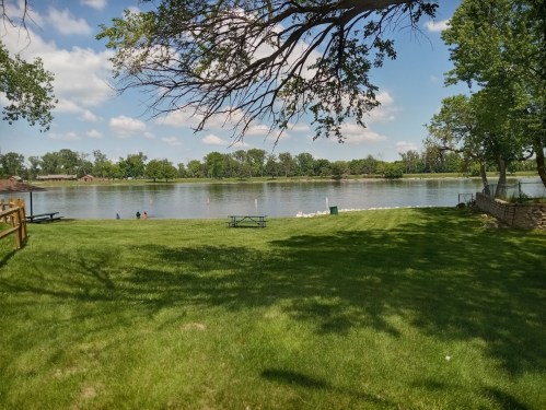 A serene lakeside view with green grass, trees, and a clear blue sky, perfect for relaxation and outdoor activities.