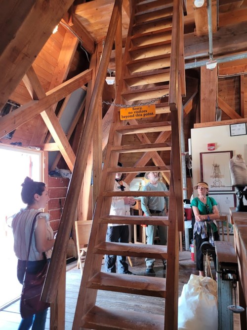 A wooden staircase leads to an upper level in a rustic building, with people chatting nearby. A sign warns not to enter.
