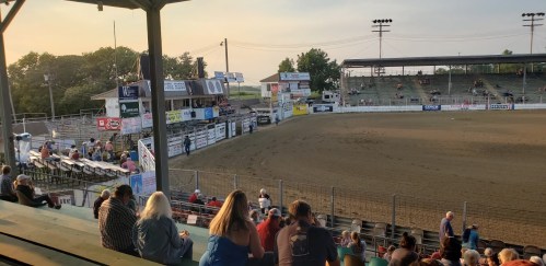 A view of a rodeo arena with empty seats and a few spectators, under a clear sky at sunset.