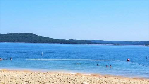 A serene beach scene with people swimming in clear blue water, surrounded by green hills under a bright blue sky.