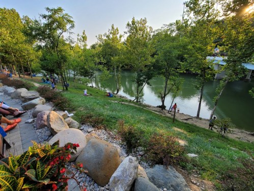 A serene riverside scene with lush greenery, rocks, and people enjoying the outdoors by the water.