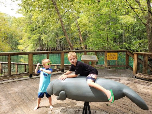 Two children play on a manatee sculpture in a wooded area, one sitting on it and the other posing beside it.