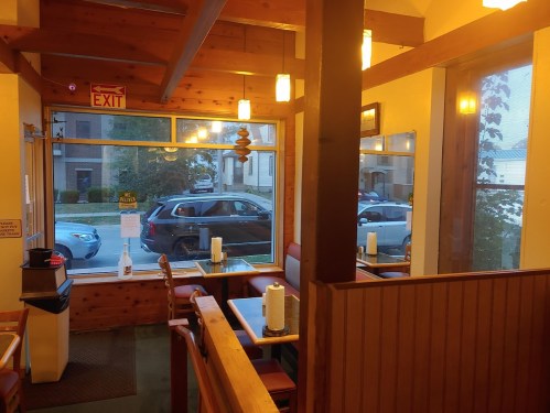Interior of a cozy restaurant with wooden beams, tables, and a view of parked cars outside through large windows.