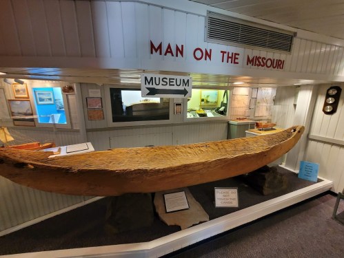 A wooden canoe displayed in a museum, with signs reading "Man on the Missouri" and "Museum" in the background.