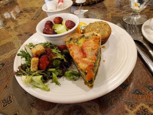 A plate with a slice of vegetable quiche, salad with croutons, a fruit bowl, and a muffin.