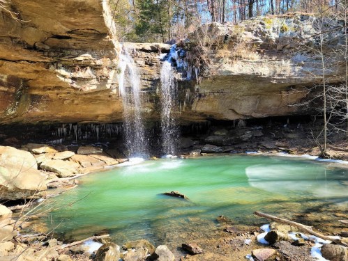 A serene waterfall cascades into a turquoise pool surrounded by rocky cliffs and winter foliage.