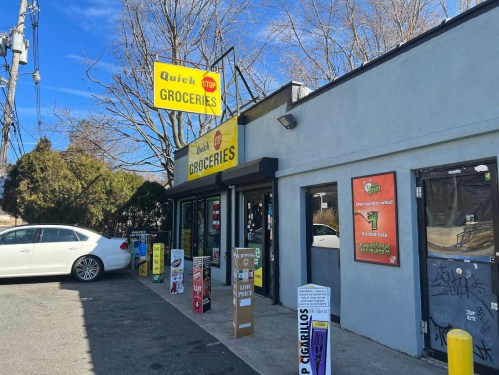 A small grocery store with a bright sign, featuring a parking area and vending machines outside.
