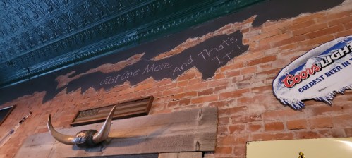 Text on a brick wall reads, "Just One More and That's It," with a sign for Coors Light nearby.