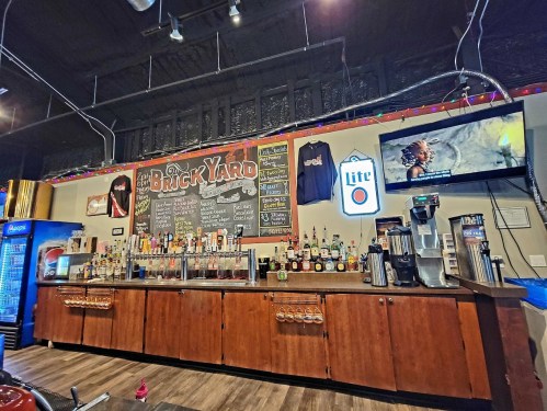 A bar with a wooden counter, shelves of liquor, a chalkboard menu, and a TV displaying a movie scene.