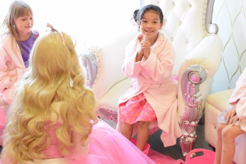 A young girl in a pink robe smiles while sitting on a chair, surrounded by friends in a bright, cheerful setting.