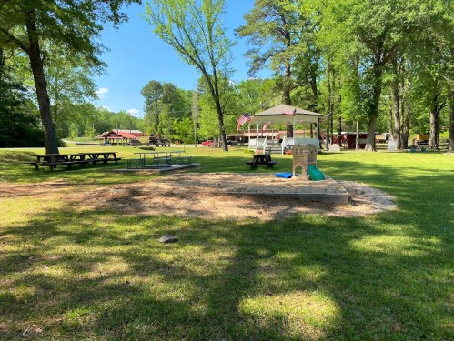 A sunny park with picnic tables, a gazebo, and green trees, featuring a sandy play area in the foreground.