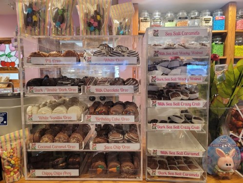Display of assorted chocolates and candies in clear containers, featuring various flavors like Oreo, caramels, and more.