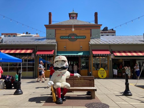 A large statue of a baseball player sits on a bench outside Findlay Market, with colorful awnings and shoppers in the background.