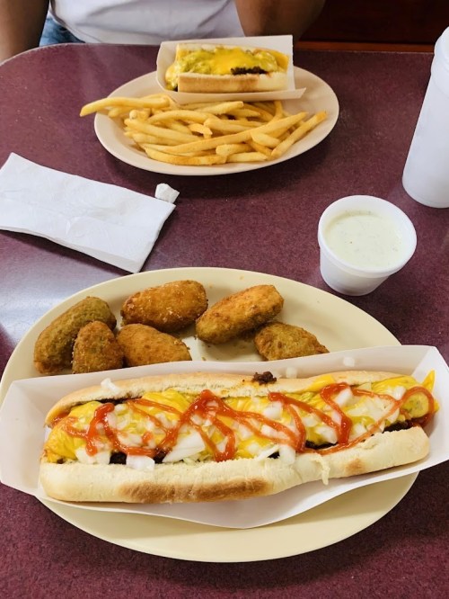A plate with a hot dog topped with mustard and ketchup, fries, and a side of fried snacks, with a drink in the background.