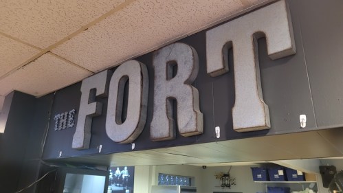 Sign reading "THE FORT" in large, metallic letters, mounted on a dark wall with hooks below.
